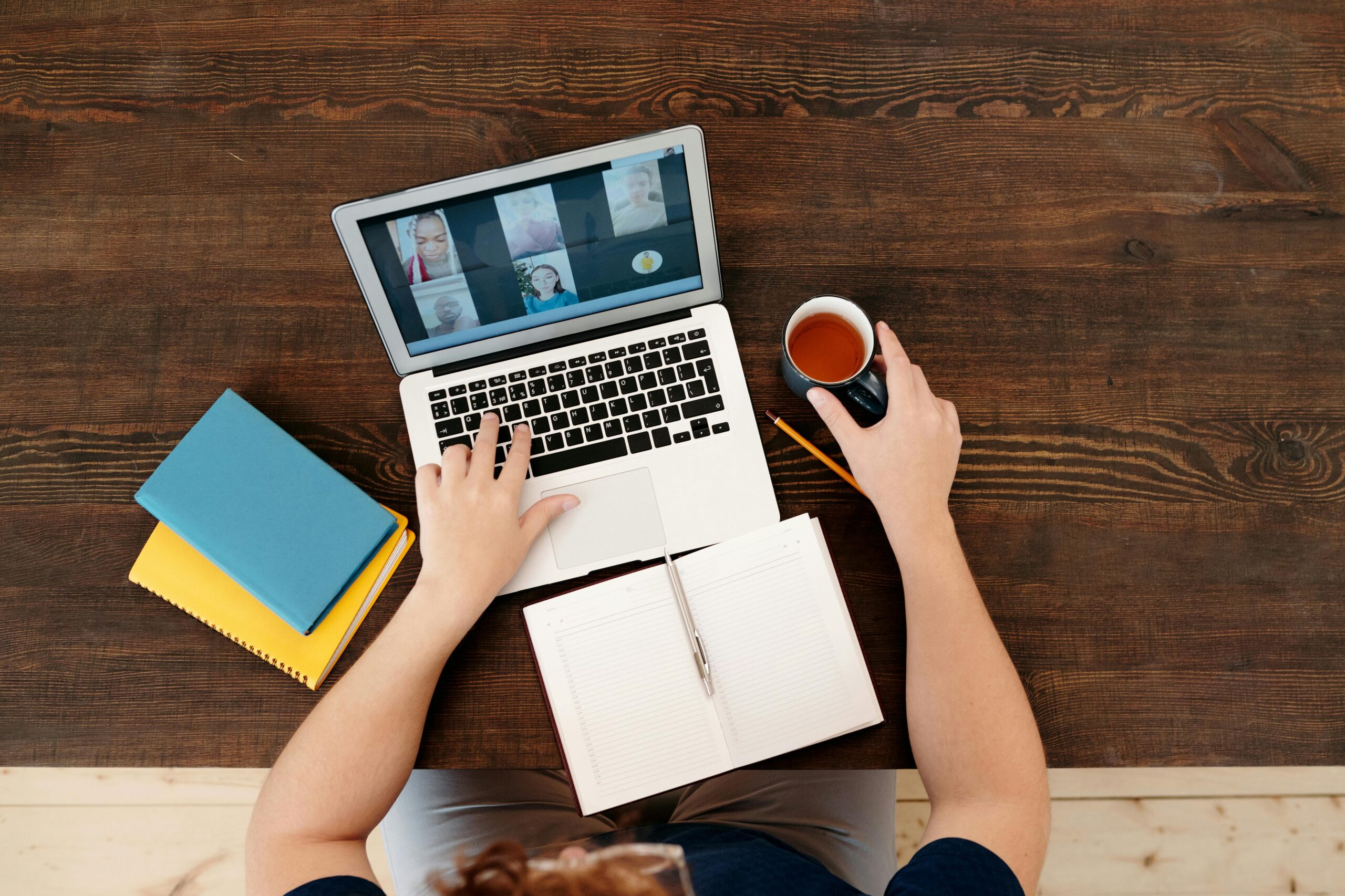 A person attending a virtual meeting on a laptop with coffee and notebooks on a wooden table.